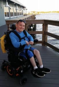 Photo of Frank Huffman, who uses a power wheelchair, smiling looking at the camera on a dock overlooking. water.